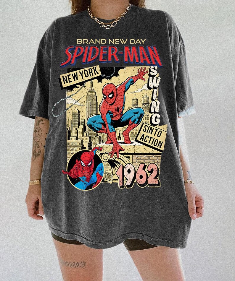 Puede incluir: Camiseta gris oscuro con un dise&ntilde;o gr&aacute;fico vintage de Spider-Man. El dise&ntilde;o incluye las palabras "BRAND NEW DAY", "SPIDER-MAN", "NEW YORK" y "1962". El superh&eacute;roe se representa en una pose din&aacute;mica sobre un fondo de paisaje urbano.