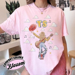 May include: A light pink t-shirt featuring a graphic illustration of a person with long hair wearing a yellow plaid shirt and cow-print bell-bottom jeans, holding a guitar and a red hat. The design includes the yellow text TS, clouds, and music notes.