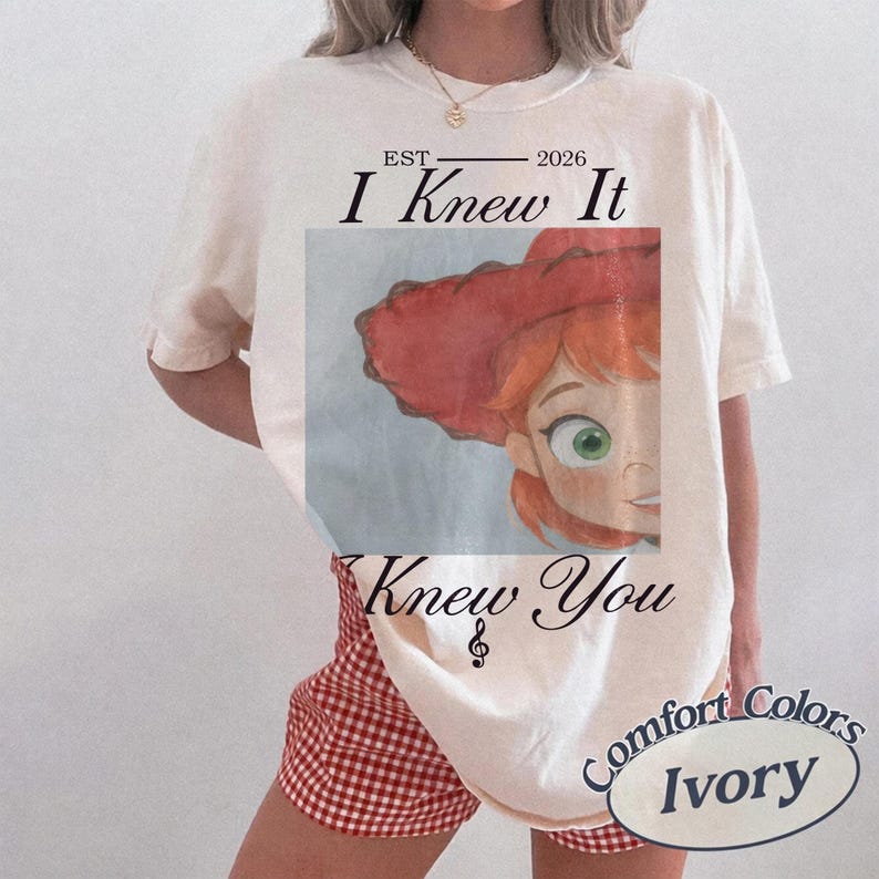 May include: An ivory short-sleeve t-shirt featuring a graphic of a cartoon character with red hair and a red hat. The text reads EST 2026 I Knew It Knew You with a treble clef symbol. The shirt is paired with red and white checkered shorts.
