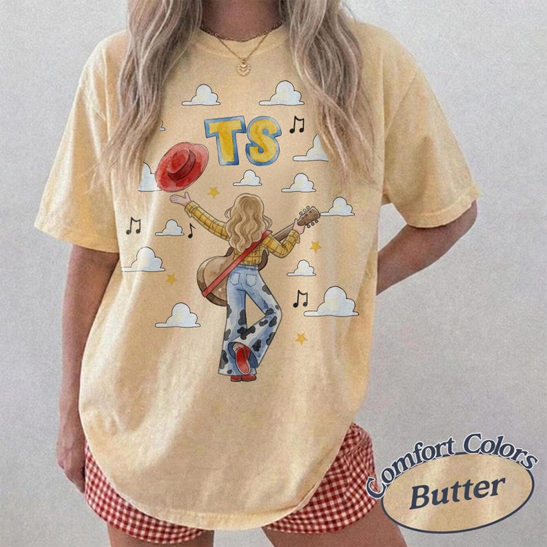 May include: A pale yellow short-sleeve t-shirt featuring a graphic print of a person with long blonde hair playing an acoustic guitar, wearing a yellow shirt and cow-print pants. The design includes the text TS, a red hat, clouds, and music notes.
