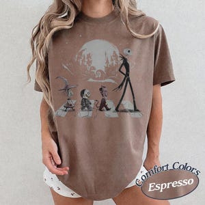May include: A brown short-sleeve t-shirt featuring a graphic print of four characters walking across a crosswalk in front of a large moon and a silhouette of a town. The design includes a witch, a skeleton, a devil, and a tall thin figure.