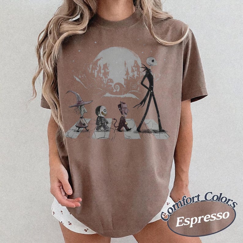 May include: A brown short-sleeve t-shirt featuring a graphic print of four characters walking across a crosswalk in front of a large moon and a silhouette of a town. The design includes a witch, a skeleton, a devil, and a tall thin figure.