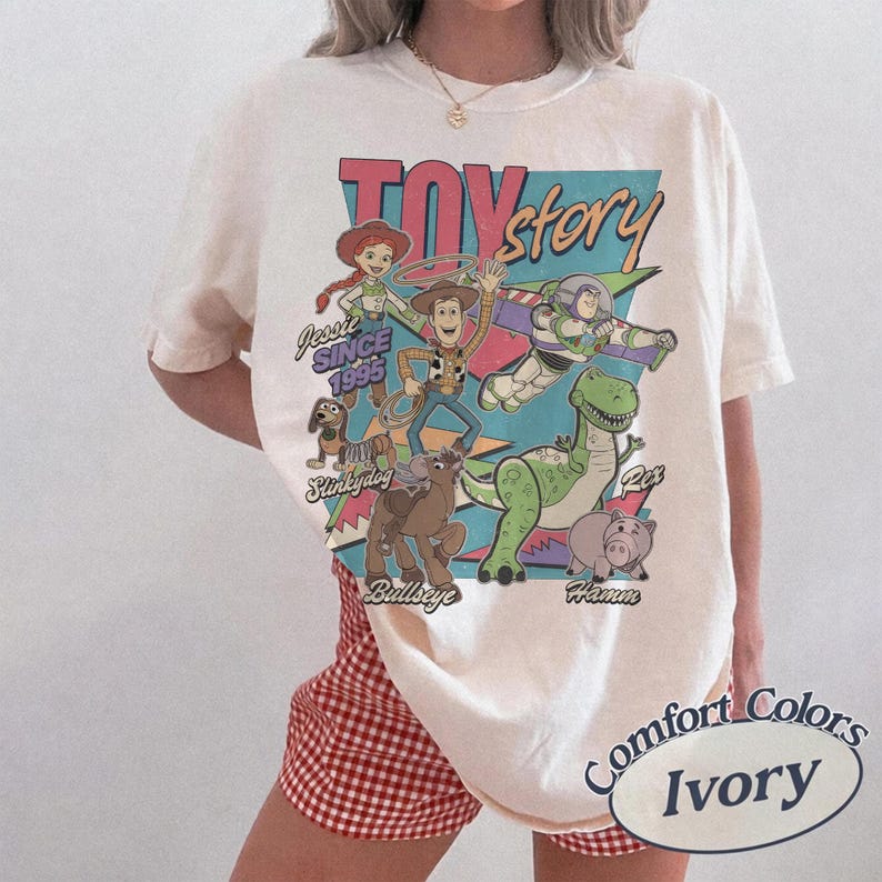 May include: Ivory-coloured t-shirt with a vibrant graphic of Toy Story characters, such as Woody and Buzz Lightyear. The design includes the text "Toy Story" and "Since 1995." The shirt is from Comfort Colors.