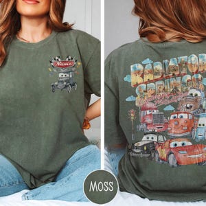 May include: A moss green t-shirt featuring a small graphic on the front chest of a cartoon car with a checkered flag and the text Name. The back displays a large graphic of multiple cartoon cars with the text Radiator Springs and Lightning McQueen.
