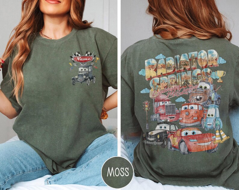 May include: A moss green t-shirt featuring a small graphic on the front chest of a cartoon car with a checkered flag and the text Name. The back displays a large graphic of multiple cartoon cars with the text Radiator Springs and Lightning McQueen.