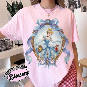 May include: Pink Comfort Colors t-shirt featuring a Cinderella graphic within an ornate gold frame with a blue ribbon. The graphic includes Cinderella in a blue gown, two mice, and candles. The text "Blossom" is printed on the bottom left.