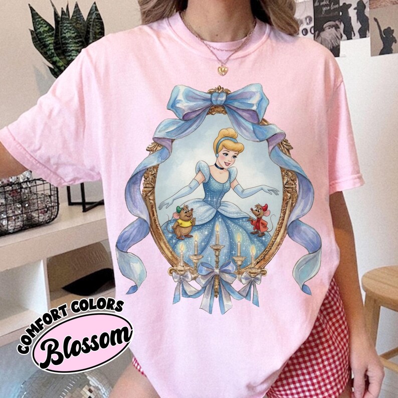May include: Pink Comfort Colors t-shirt featuring a Cinderella graphic within an ornate gold frame with a blue ribbon. The graphic includes Cinderella in a blue gown, two mice, and candles. The text "Blossom" is printed on the bottom left.