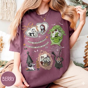 May include: Berry-colored t-shirt featuring multiple framed illustrations of characters from "The Nightmare Before Christmas." The illustrations include Jack Skellington, Sally, Oogie Boogie, and Zero. The text "The Nightmare Before Christmas" is also present.