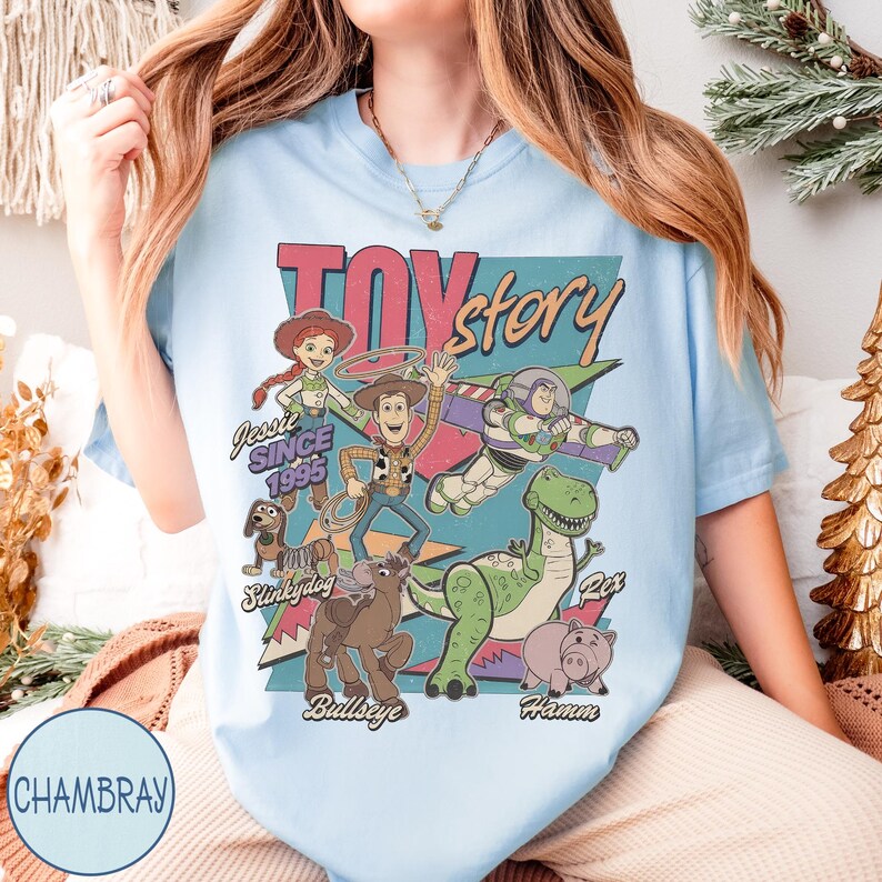 May include: Pale blue t-shirt with a retro graphic of Toy Story characters, including Woody, Buzz Lightyear, and Jessie. The text "Toy Story" and "Since 1995" are also displayed. The design has a vintage style.