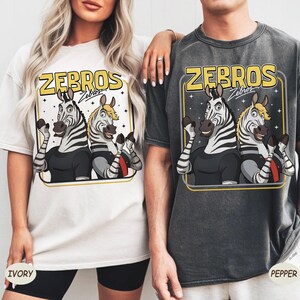 May include: Ivory and pepper-coloured t-shirts featuring a cartoon graphic of two zebras with the text "ZEBROS". The zebras are wearing black shirts and waving. The ivory shirt has the word "IVORY" printed on it.
