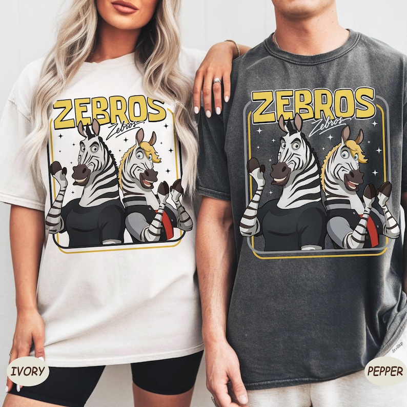 May include: Ivory and pepper-coloured t-shirts featuring a cartoon graphic of two zebras with the text "ZEBROS". The zebras are wearing black shirts and waving. The ivory shirt has the word "IVORY" printed on it.