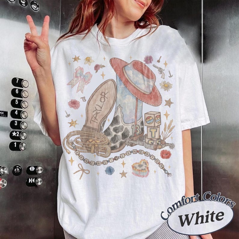 May include: A white t-shirt featuring a graphic print of a cowboy boot, a hat, a lasso, and blocks with the letter T. The design includes the name Taylor, the number 13, and various small illustrations like stars, flowers, and bows in soft colors.