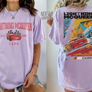 May include: Lavender t-shirt featuring Lightning McQueen from the Cars movie. The front graphic includes the text "Lightning McQueen" and "Cars", with a red race car and checkered flags. The back displays a larger graphic of McQueen racing.