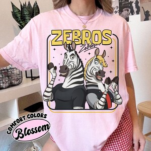 May include: Pale pink t-shirt featuring a cartoon illustration of two zebras. The zebras are dressed in black and grey outfits, within a yellow and pink frame, with the word "ZEBROS". The shirt also has the words "Comfort Colors" and "Blossom".