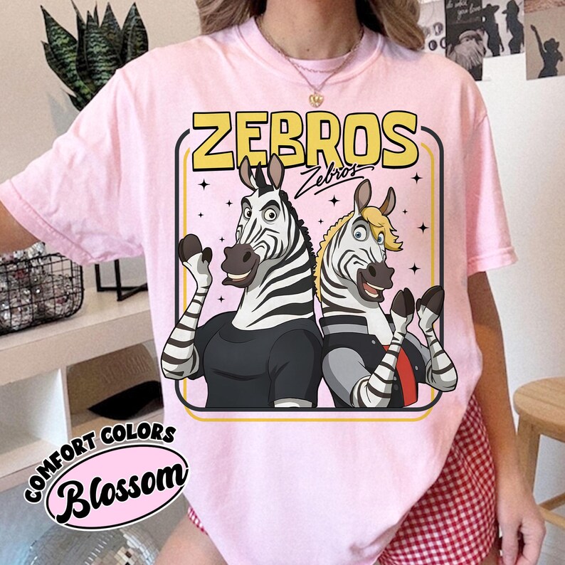 May include: Pale pink t-shirt featuring a cartoon illustration of two zebras. The zebras are dressed in black and grey outfits, within a yellow and pink frame, with the word "ZEBROS". The shirt also has the words "Comfort Colors" and "Blossom".