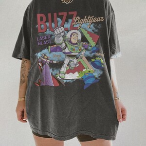 May include: Dark grey oversized t-shirt with a graphic of Buzz Lightyear and the text "BUZZ Lightyear" and "LASER BLAST". The design includes other characters and a colourful background.