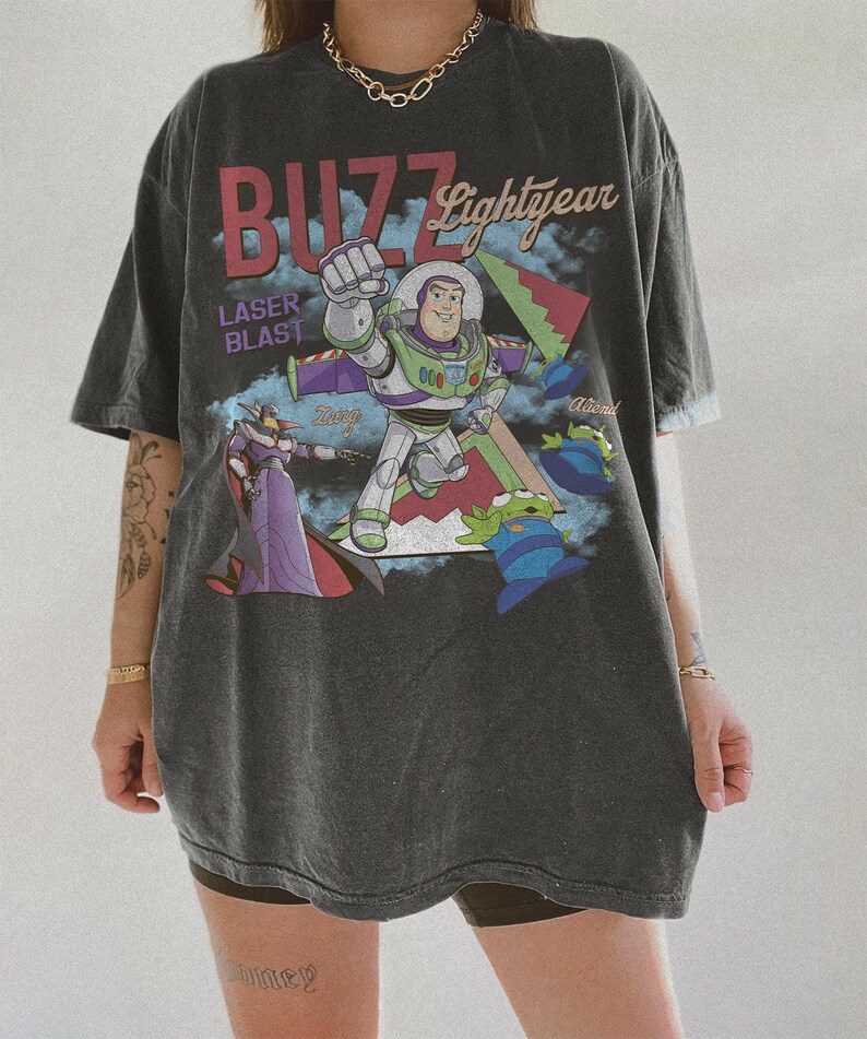 May include: Dark grey oversized t-shirt with a graphic of Buzz Lightyear and the text "BUZZ Lightyear" and "LASER BLAST". The design includes other characters and a colourful background.