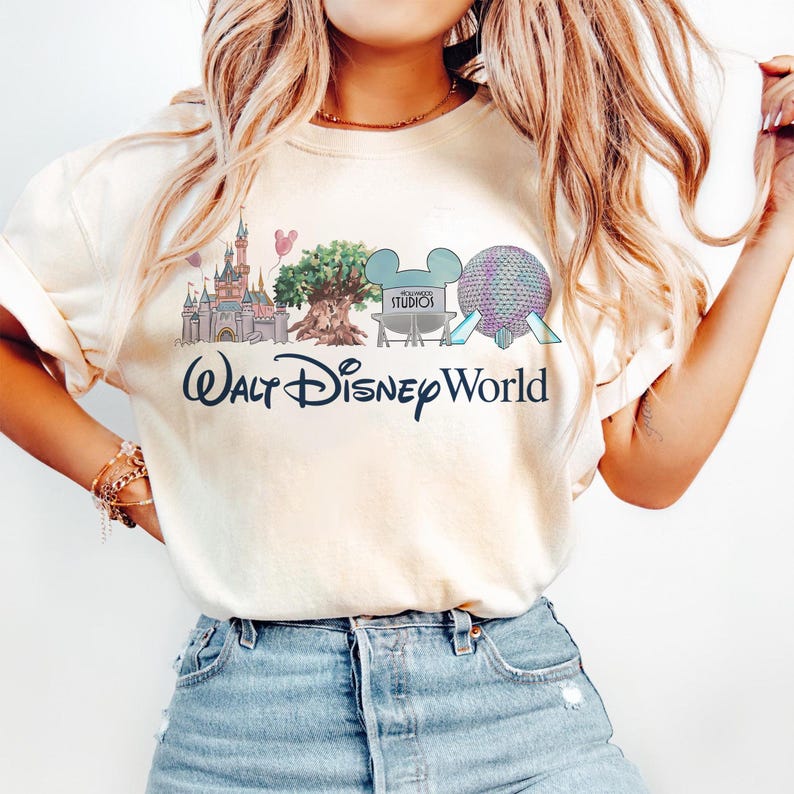 May include: Cream-colored t-shirt featuring a Walt Disney World graphic. The design includes illustrations of Cinderella's castle, the Tree of Life, a Mickey Mouse head with "Studios" text, and Spaceship Earth. The words "Walt Disney World" are written in a script font.
