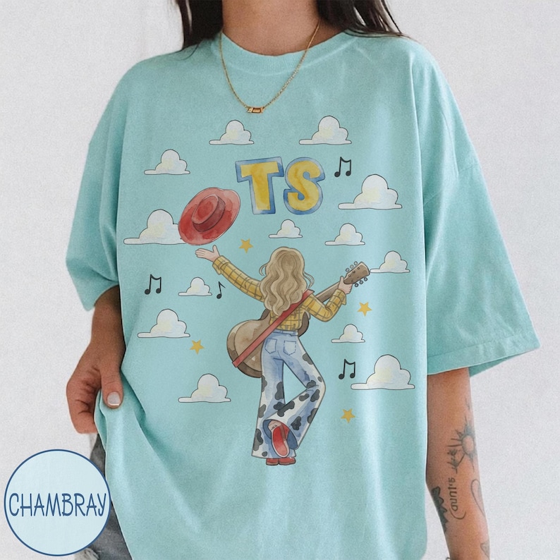 May include: A light blue t-shirt featuring a graphic of a person with long hair wearing a yellow plaid shirt and cow-print bell-bottom pants. The person holds a guitar and a red hat. The design includes the letters TS, clouds, stars, and music notes.