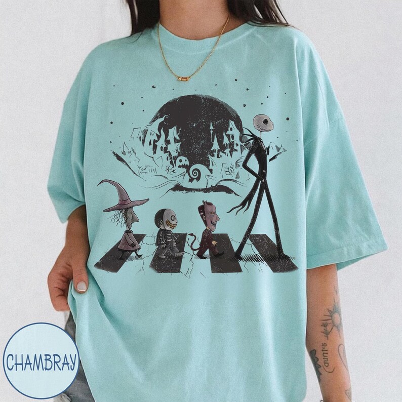 May include: A light blue t-shirt featuring a black graphic print of characters from The Nightmare Before Christmas walking across a crosswalk. The design includes a large moon, a spooky house silhouette, and the characters Lock, Shock, and Barrel.