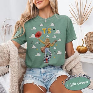 May include: A sage green short-sleeve t-shirt featuring a graphic of a person with long hair holding a guitar and a red hat. The design includes white clouds, yellow stars, musical notes, and the letters TS in yellow and blue against a sky background.