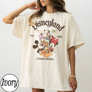 Puede incluir: Camiseta de gran tama&ntilde;o de color marfil con un gr&aacute;fico de Disneyland. El gr&aacute;fico incluye a Mickey Mouse, Minnie Mouse, Donald Duck, Daisy Duck, Goofy y Pluto. El texto "Disneyland" est&aacute; arqueado en la parte superior, con "Est 1955" y "California" debajo.