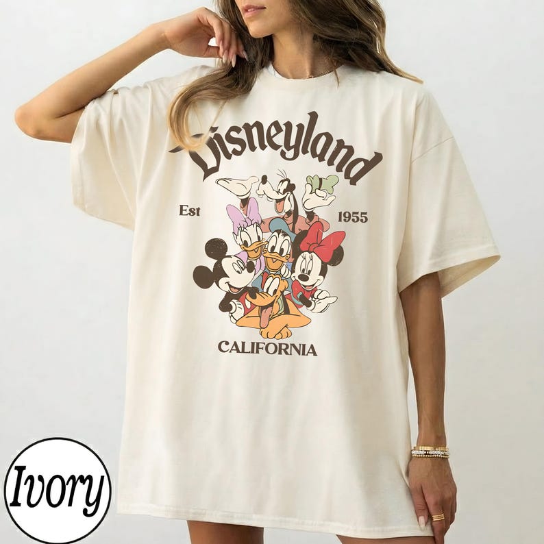 Puede incluir: Camiseta de gran tama&ntilde;o de color marfil con un gr&aacute;fico de Disneyland. El gr&aacute;fico incluye a Mickey Mouse, Minnie Mouse, Donald Duck, Daisy Duck, Goofy y Pluto. El texto "Disneyland" est&aacute; arqueado en la parte superior, con "Est 1955" y "California" debajo.