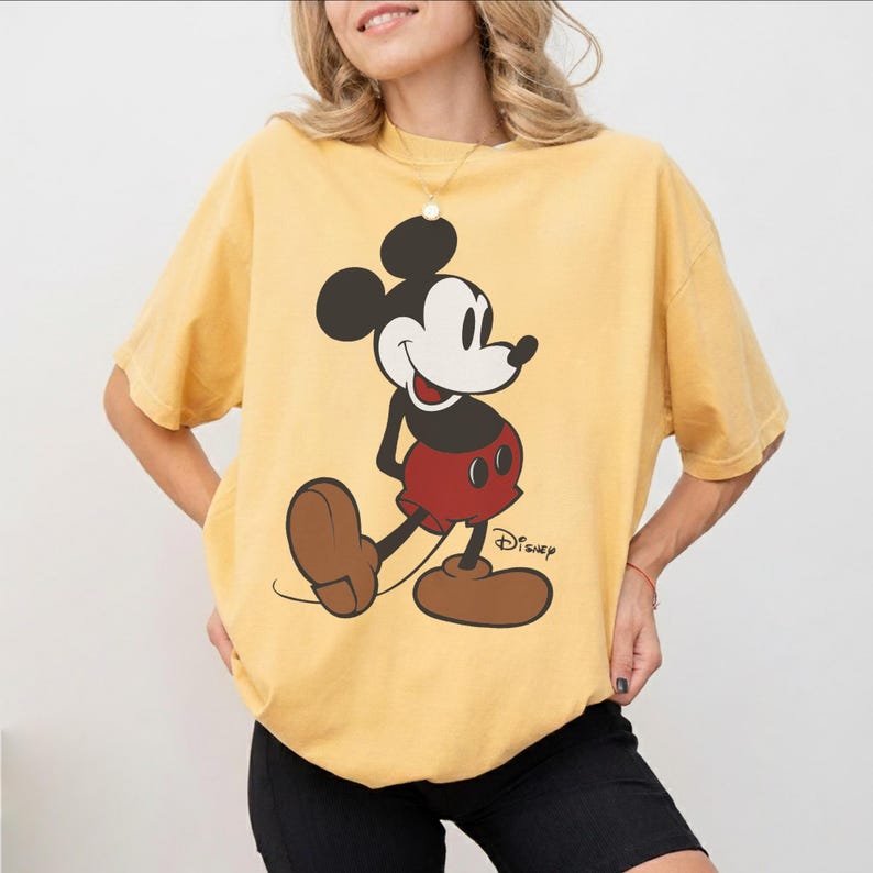 May include: Yellow t-shirt featuring a classic Mickey Mouse graphic. The cartoon character is depicted in black, white, red, and brown, with the Disney logo printed below. The shirt has short sleeves and a relaxed fit.