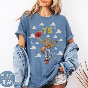 May include: A blue t-shirt featuring a graphic of a person with long hair wearing a yellow plaid shirt and cow-print bell-bottom pants, holding a guitar and a red hat. The design includes the text TS, musical notes, stars, and white clouds.