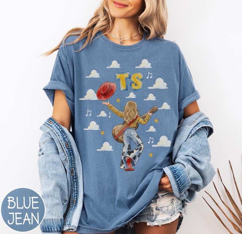 May include: A blue t-shirt featuring a graphic of a person with long hair wearing a yellow plaid shirt and cow-print bell-bottom pants, holding a guitar and a red hat. The design includes the text TS, musical notes, stars, and white clouds.