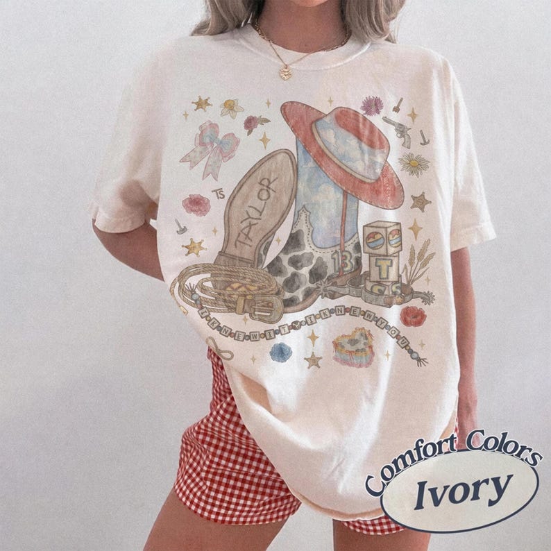 May include: An ivory t-shirt featuring a graphic print of a cowboy boot, a red-brimmed hat, a lasso, a bow, and a block with the letter T. The boot is marked with the name Taylor and the number 13. The design includes stars, flowers, and text.