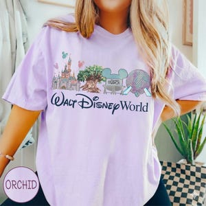 May include: A lilac t-shirt with a graphic of Walt Disney World landmarks, including a castle, tree, and Spaceship Earth. The words "Walt Disney World" are printed in a decorative font. The shirt has short sleeves.