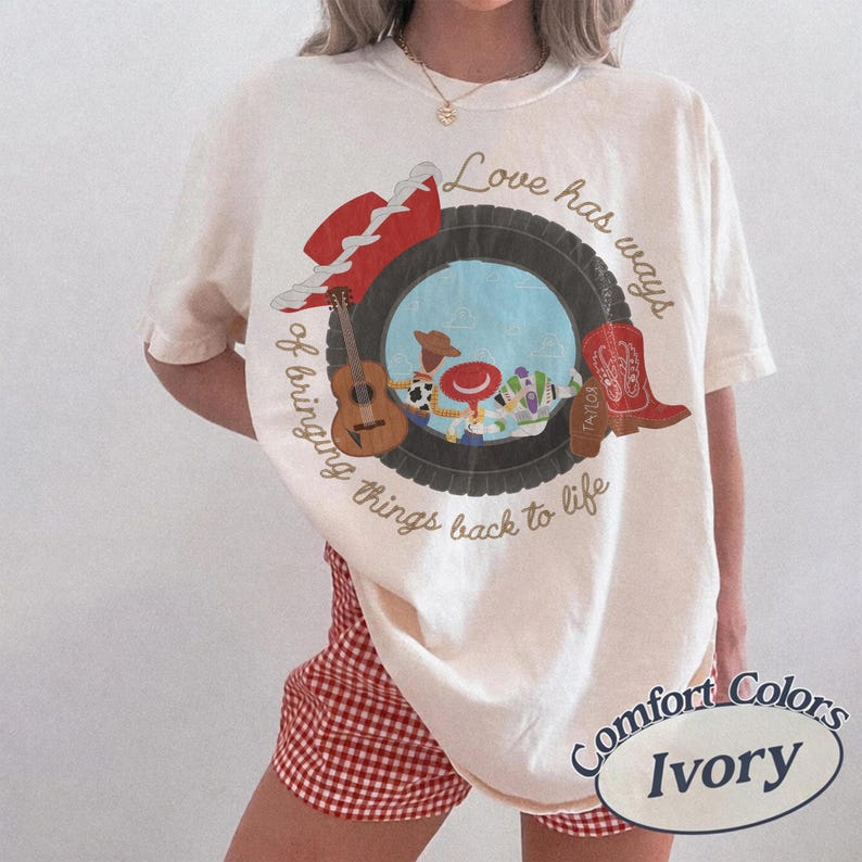 K&ouml;nnte beinhalten: Ein elfenbeinfarbenes Kurzarm-T-Shirt mit einer Grafik, die einen Reifen zeigt, der Zeichentrickfiguren, eine Gitarre, einen roten Hut und einen roten Cowboystiefel umrahmt. Der Text lautet, Love has ways of bringing things back to life.