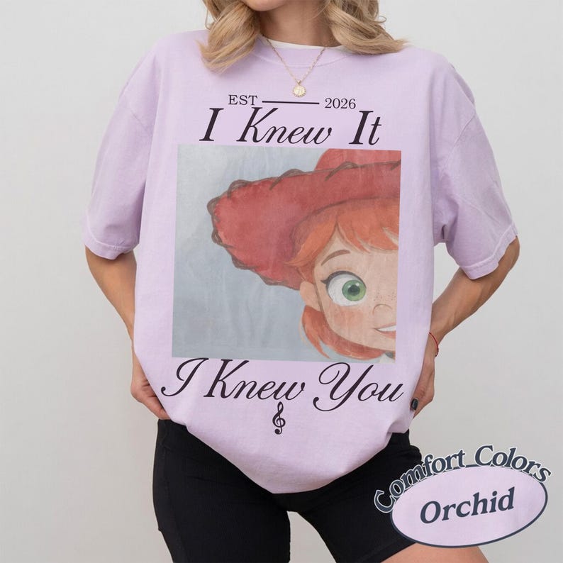May include: A light purple t-shirt featuring a graphic print of a cartoon character with red hair and a red hat. The text reads EST 2026 I Knew It I Knew You with a musical treble clef symbol below the text. The shirt has a crew neckline and short sleeves.