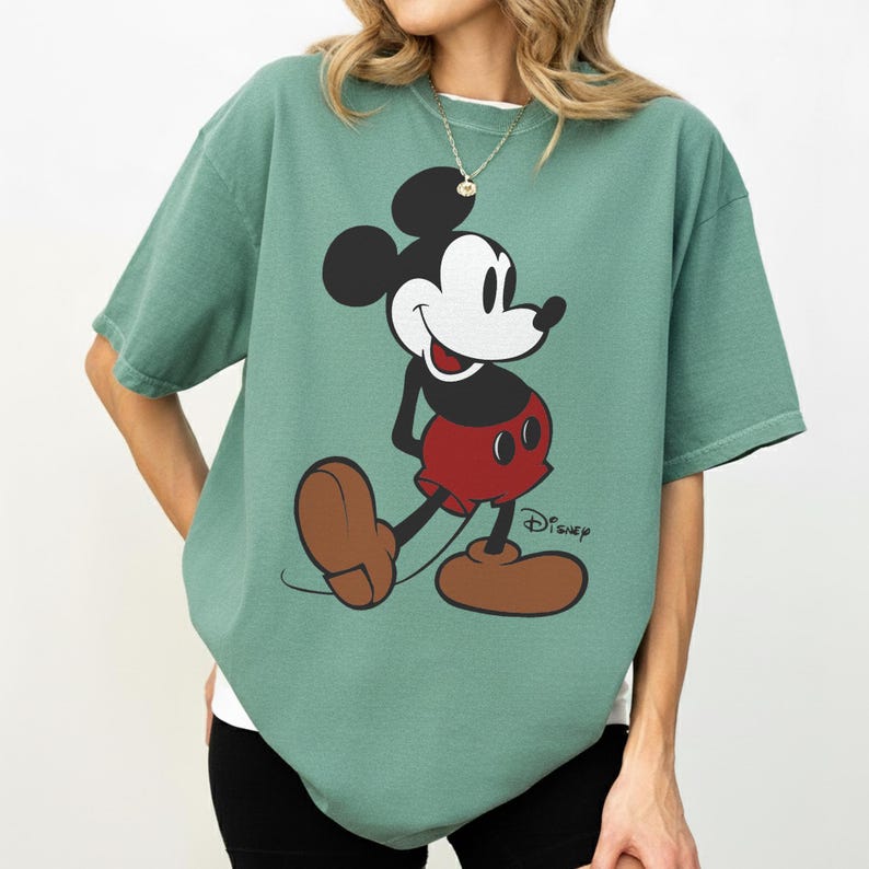 May include: Sage green t-shirt featuring a graphic of Mickey Mouse in his classic pose. Mickey is depicted in black, white, red, and brown. The Disney logo is printed in black on the lower right of the shirt.