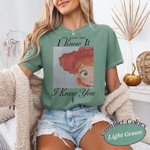 May include: A light green short-sleeve t-shirt featuring a graphic of a cartoon character with red hair and a red hat. The text reads EST 2026 I Knew It I Knew You with a small musical note symbol below the text, centered on the chest.