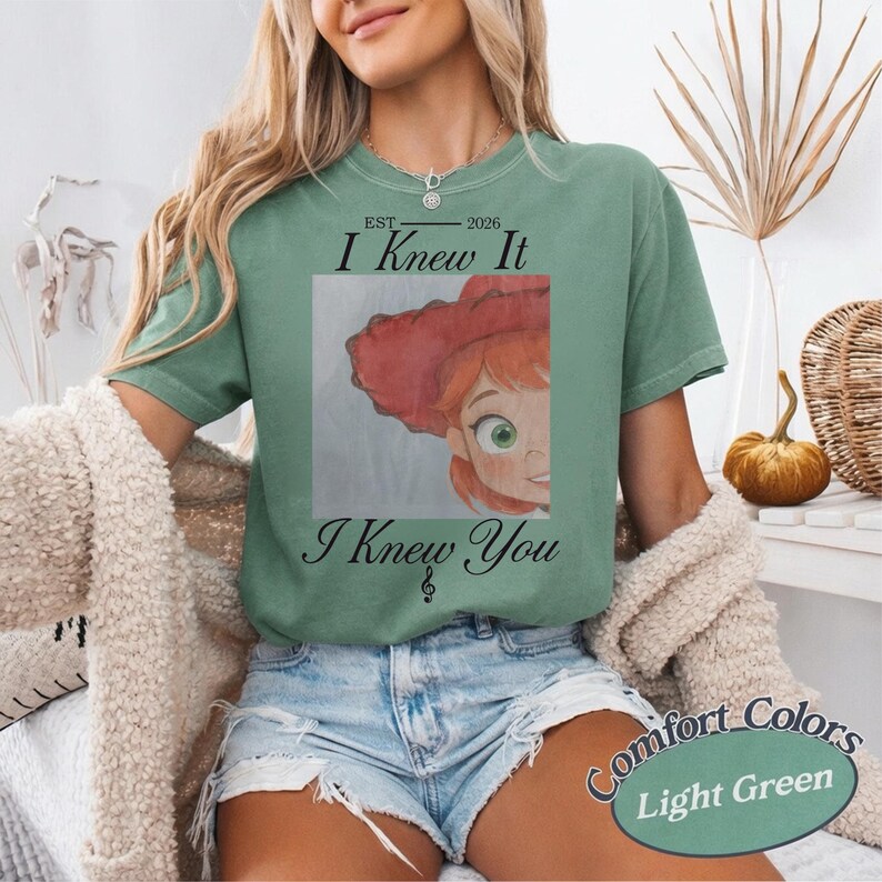 May include: A light green short-sleeve t-shirt featuring a graphic of a cartoon character with red hair and a red hat. The text reads EST 2026 I Knew It I Knew You with a small musical note symbol below the text, centered on the chest.
