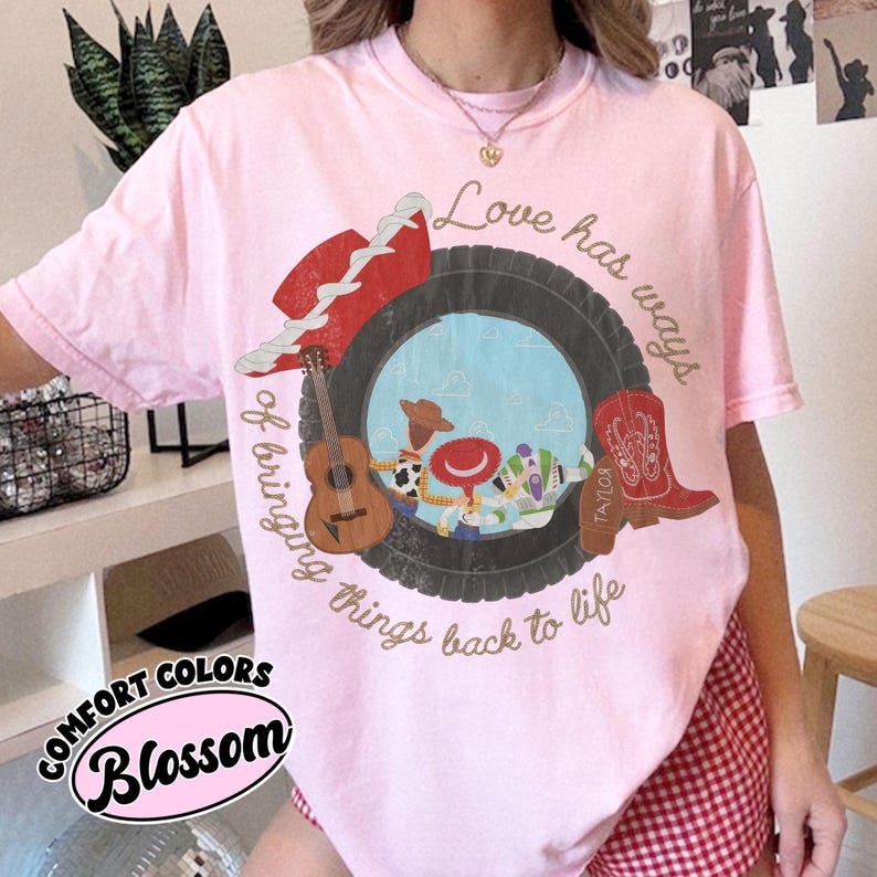 K&ouml;nnte beinhalten: Ein hellrosa T-Shirt mit einer Grafik aus einem schwarzen Reifen, der Zeichentrickfiguren, eine braune Akustikgitarre, einen roten Cowboyhut und einen roten Cowboystiefel umrahmt. Der Text lautet Love has ways of bringing things back to life.