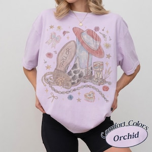 May include: A light purple short-sleeve t-shirt featuring a graphic print of a cowboy boot, a hat, a belt, and building blocks. The design includes the text Taylor and 13, surrounded by small stars, flowers, and a bow. The shirt has a crew neckline.