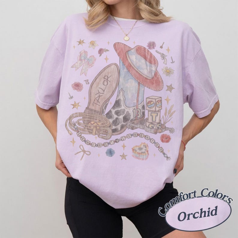May include: A light purple short-sleeve t-shirt featuring a graphic print of a cowboy boot, a hat, a belt, and building blocks. The design includes the text Taylor and 13, surrounded by small stars, flowers, and a bow. The shirt has a crew neckline.