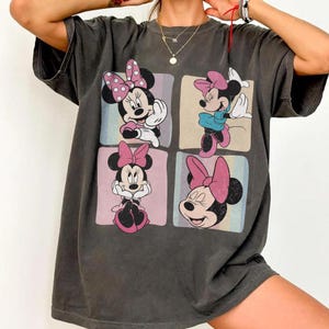Op de afbeelding: Donkergrijs T-shirt met vier panelen met Minnie Mouse-illustraties. Elk paneel toont een andere pose van Minnie Mouse, allemaal met roze strikken. De achtergrond van elk paneel heeft pastelkleurige strepen.