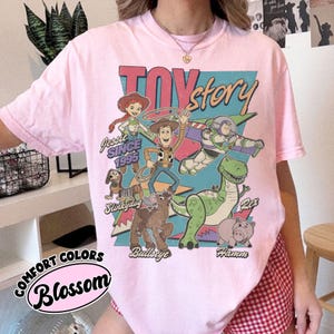 May include: Pale pink t-shirt with a graphic of "Toy Story" characters, including Woody, Buzz Lightyear, Jessie, Rex, Hamm, Slinky Dog, and Bullseye. The design also features the words "Toy Story" and "Since 1995."