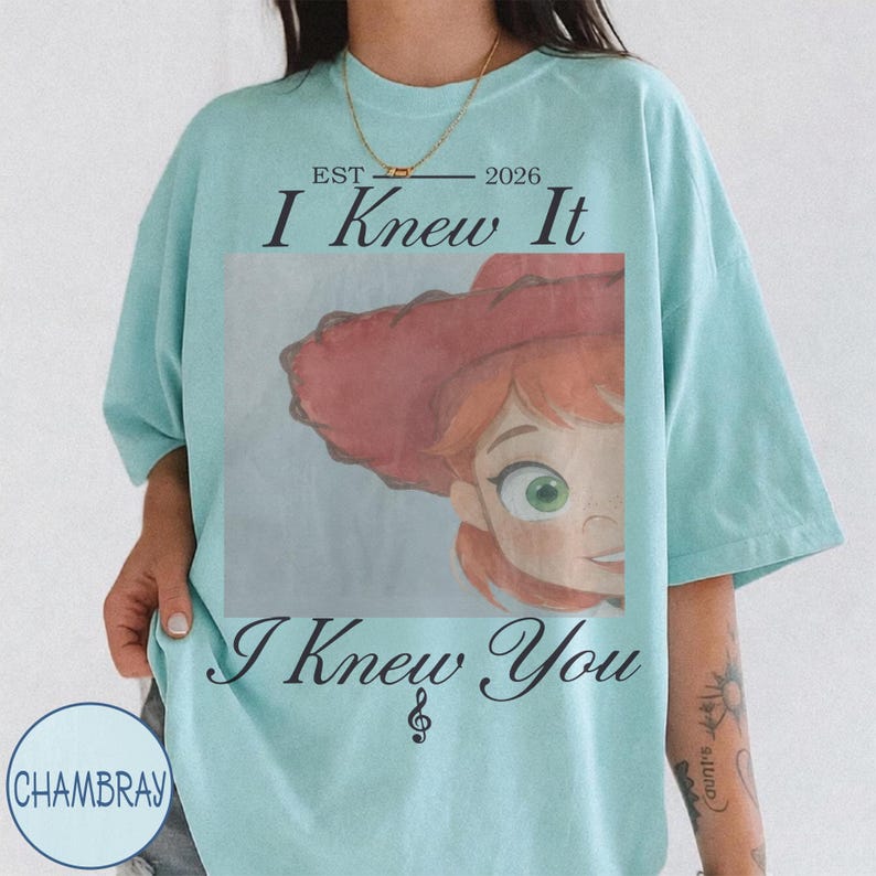 May include: A light blue t-shirt featuring a graphic of a character with red hair and a red hat. The text reads EST 2026 I Knew It I Knew You with a musical note symbol. The shirt has short sleeves and a crew neckline, displaying a casual fit.