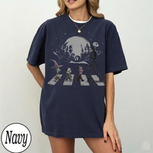 May include: A navy blue short-sleeve t-shirt featuring a graphic print of characters from The Nightmare Before Christmas walking across a crosswalk. The design includes a large moon, a spooky town silhouette, and a circular navy color label.