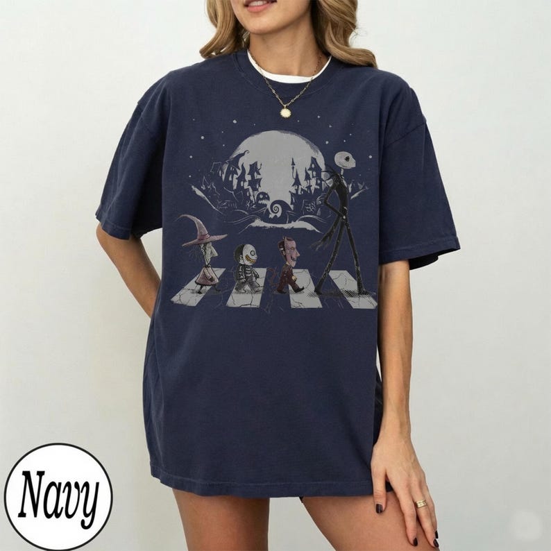 May include: A navy blue short-sleeve t-shirt featuring a graphic print of characters from The Nightmare Before Christmas walking across a crosswalk. The design includes a large moon, a spooky town silhouette, and a circular navy color label.
