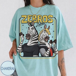 May include: A pale blue t-shirt with a cartoon illustration of two zebras, each wearing a shirt, with the text "ZEBROS". The zebras are in a retro style and have their arms raised. The t-shirt has a loose fit.