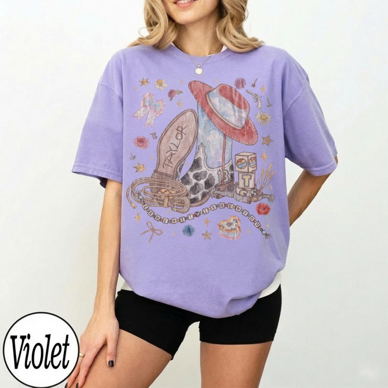 May include: A light purple short-sleeve t-shirt featuring a graphic print of a cowboy boot, a hat, a lasso, and blocks with the text "13" and "T". The design includes small stars, flowers, and a beaded string with the text "TAYLOR" on the boot.