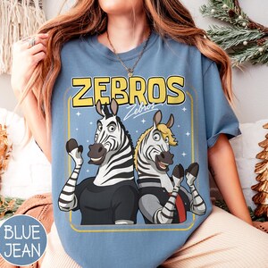 May include: Blue jean coloured t-shirt with a cartoon graphic of two zebras and the yellow text "ZEBROS". The zebras are wearing black and grey outfits and waving. The shirt has a crew neck.