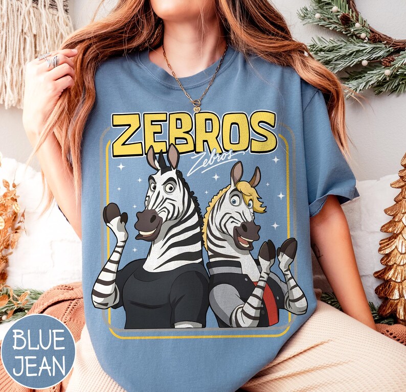 May include: Blue jean coloured t-shirt with a cartoon graphic of two zebras and the yellow text "ZEBROS". The zebras are wearing black and grey outfits and waving. The shirt has a crew neck.