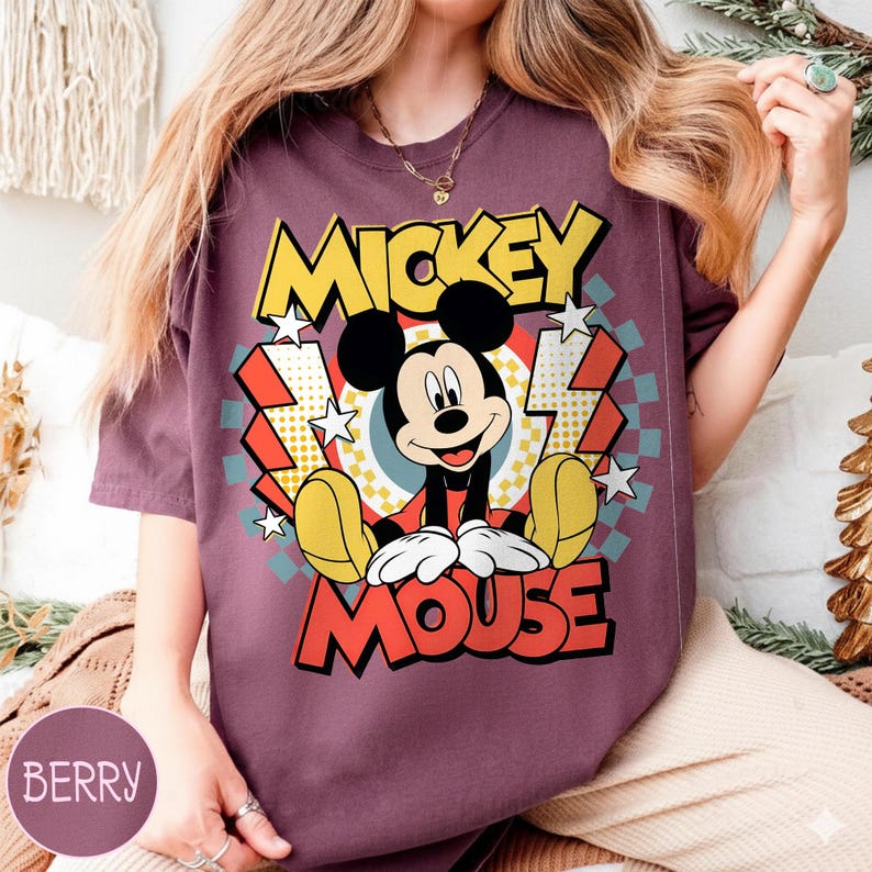 May include: Berry-colored t-shirt featuring a graphic of Mickey Mouse. The design includes the words "Mickey Mouse" in yellow and red, with Mickey Mouse sitting in the center. The background has a retro design with stars and lightning bolts.